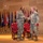 Two U.S. Army soldiers in Operational Camouflage Pattern (OCP) uniforms are shaking hands on a formal stage, suggesting a military ceremony such as a promotion, award presentation, or change of command. The setting includes red leather chairs and a row of state and military flags displayed on a wooden-paneled backdrop. The formal staging and flag arrangement indicate an official military function.