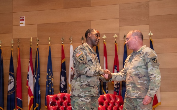 Two U.S. Army soldiers in Operational Camouflage Pattern (OCP) uniforms are shaking hands on a formal stage, suggesting a military ceremony such as a promotion, award presentation, or change of command. The setting includes red leather chairs and a row of state and military flags displayed on a wooden-paneled backdrop. The formal staging and flag arrangement indicate an official military function.