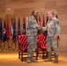 Two U.S. Army soldiers in Operational Camouflage Pattern (OCP) uniforms are shaking hands on a formal stage, suggesting a military ceremony such as a promotion, award presentation, or change of command. The setting includes red leather chairs and a row of state and military flags displayed on a wooden-paneled backdrop. The formal staging and flag arrangement indicate an official military function.