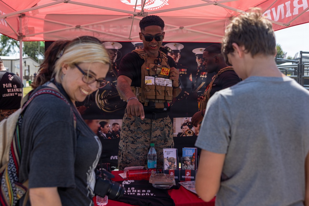 A U.S. Marines recruiting booth at what appears to be an outdoor public event or fair. A Marine in camouflage uniform and tactical vest is engaging with civilian visitors at a table displaying recruitment materials. The atmosphere is friendly and interactive, with the Marine smiling and presenting items to attendees.
