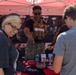 A U.S. Marines recruiting booth at what appears to be an outdoor public event or fair. A Marine in camouflage uniform and tactical vest is engaging with civilian visitors at a table displaying recruitment materials. The atmosphere is friendly and interactive, with the Marine smiling and presenting items to attendees.