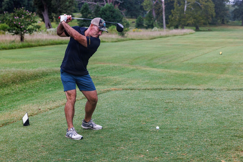 A middle-aged man is captured mid-swing on a golf course tee box, having just completed or nearly completed a driver swing. The golf ball remains on the tee, suggesting the photo was taken at the moment of impact or just before. The setting is a well-maintained golf course with lush fairway, rough areas, and tree-lined background.