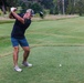 A middle-aged man is captured mid-swing on a golf course tee box, having just completed or nearly completed a driver swing. The golf ball remains on the tee, suggesting the photo was taken at the moment of impact or just before. The setting is a well-maintained golf course with lush fairway, rough areas, and tree-lined background.