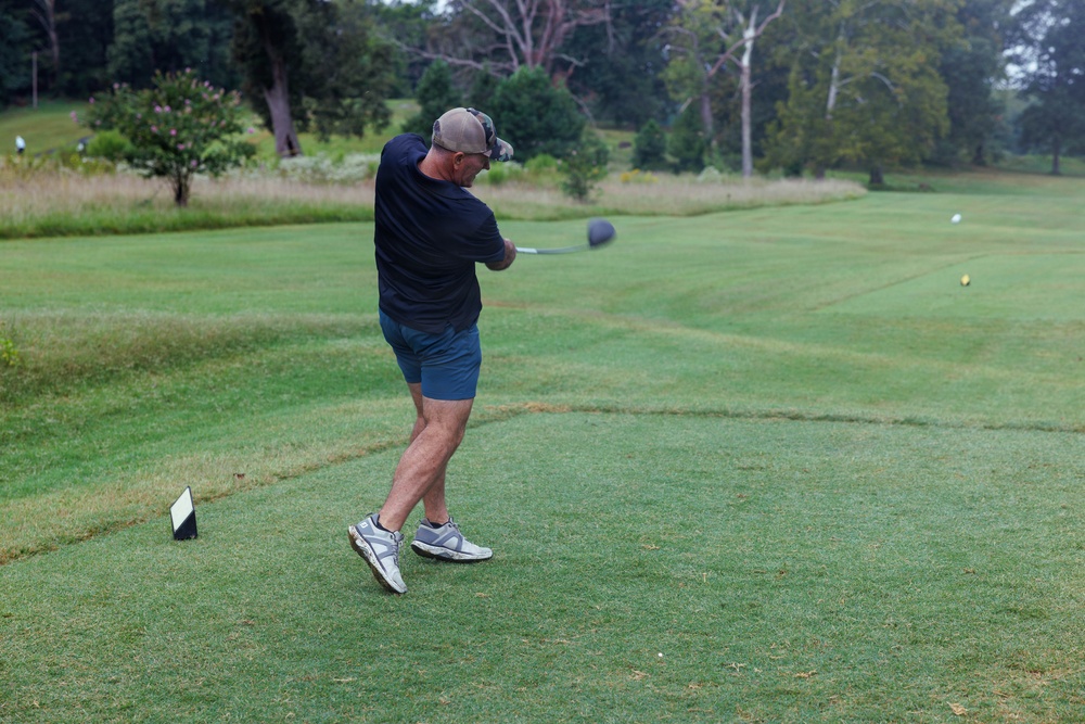 A middle-aged man is captured mid-swing on a golf course tee box, having just completed a driver swing. He is dressed casually in a dark navy shirt, blue shorts, and athletic sneakers with a baseball cap. The well-maintained golf course stretches into the background with trees and rough grass visible.