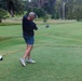 A middle-aged man is captured mid-swing on a golf course tee box, having just completed a driver swing. He is dressed casually in a dark navy shirt, blue shorts, and athletic sneakers with a baseball cap. The well-maintained golf course stretches into the background with trees and rough grass visible.
