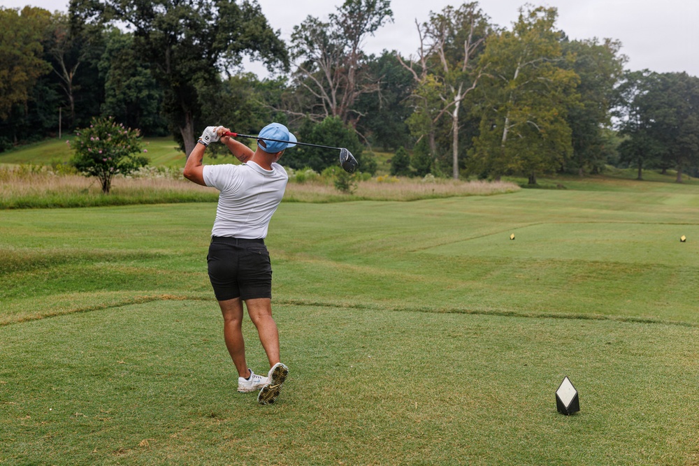 A golfer is captured mid-swing on a golf course tee box, having just completed a drive shot. The player is facing away from the camera, wearing casual golf attire on a lush green fairway surrounded by trees. The scene appears to be taken on an overcast day at a well-maintained golf course.