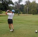 A golfer is captured mid-swing on a golf course tee box, having just completed a drive shot. The player is facing away from the camera, wearing casual golf attire on a lush green fairway surrounded by trees. The scene appears to be taken on an overcast day at a well-maintained golf course.