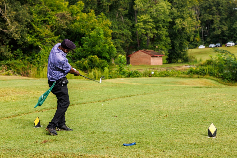 A golfer is captured mid-swing on a golf course tee box, having just struck the ball which is visible in the air downrange. The player is wearing a purple/blue shirt, black pants, and a dark cap, with a green towel hanging from their belt. The course features lush green fairway, surrounding trees, a brick building in the background, and parked vehicles on an elevated area.