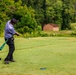 A golfer is captured mid-swing on a golf course tee box, having just struck the ball which is visible in the air downrange. The player is wearing a purple/blue shirt, black pants, and a dark cap, with a green towel hanging from their belt. The course features lush green fairway, surrounding trees, a brick building in the background, and parked vehicles on an elevated area.