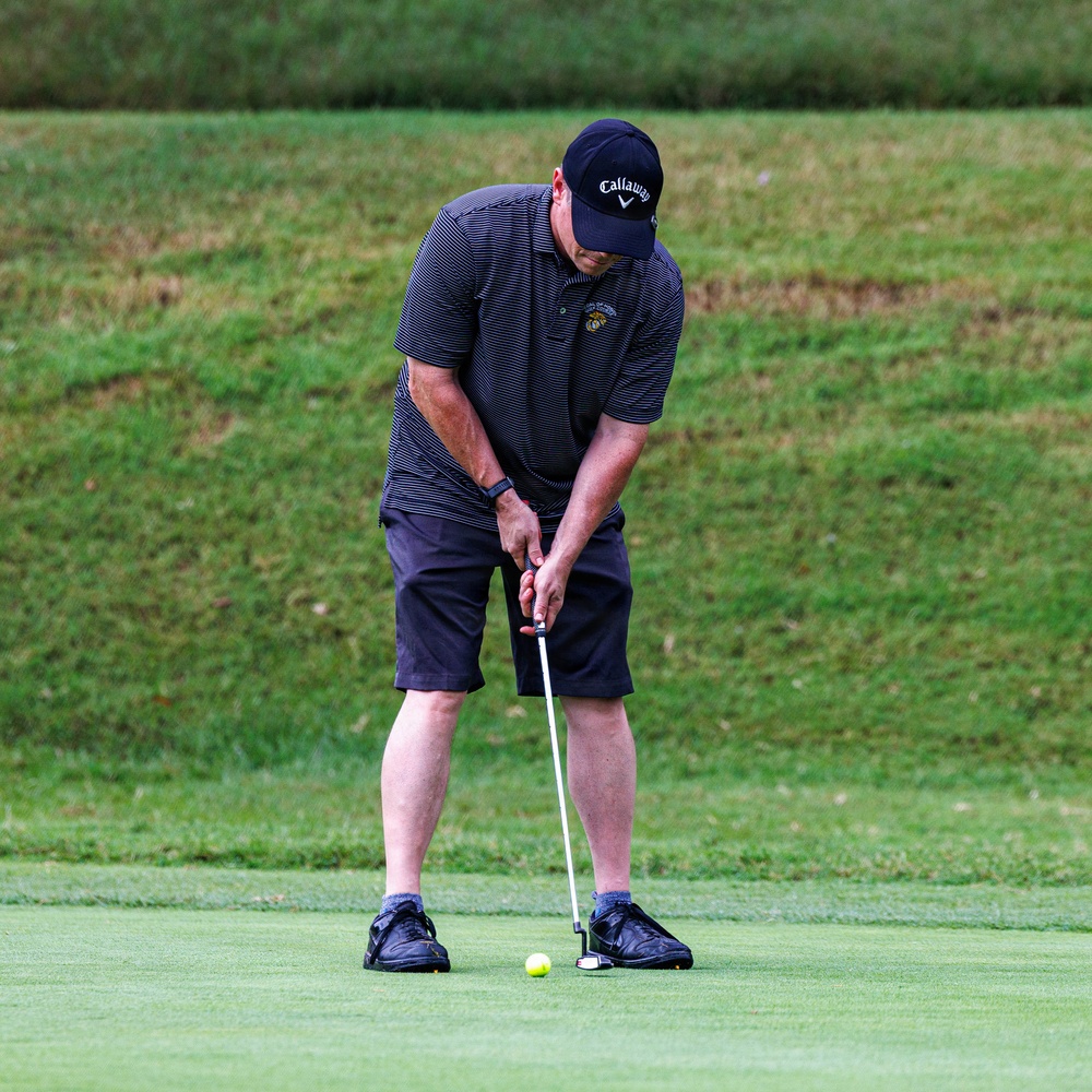 A man is putting on a golf green, positioned over a yellow golf ball with a putter. He is wearing a Callaway-branded cap, a dark striped polo shirt with a small emblem on the chest, dark shorts, and black athletic shoes. The background shows a grassy hillside typical of a golf course.