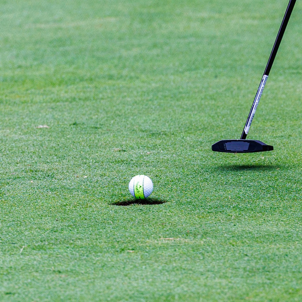 A close-up shot of a golf putting green showing a white Callaway golf ball positioned at the edge of the cup hole. A black putter club head with a dark shaft is visible to the right, positioned as if about to make or having just made a putt. The well-manicured green grass is in sharp focus, suggesting a professional or well-maintained golf course.