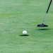 A close-up shot of a golf putting green showing a white Callaway golf ball positioned at the edge of the cup hole. A black putter club head with a dark shaft is visible to the right, positioned as if about to make or having just made a putt. The well-manicured green grass is in sharp focus, suggesting a professional or well-maintained golf course.