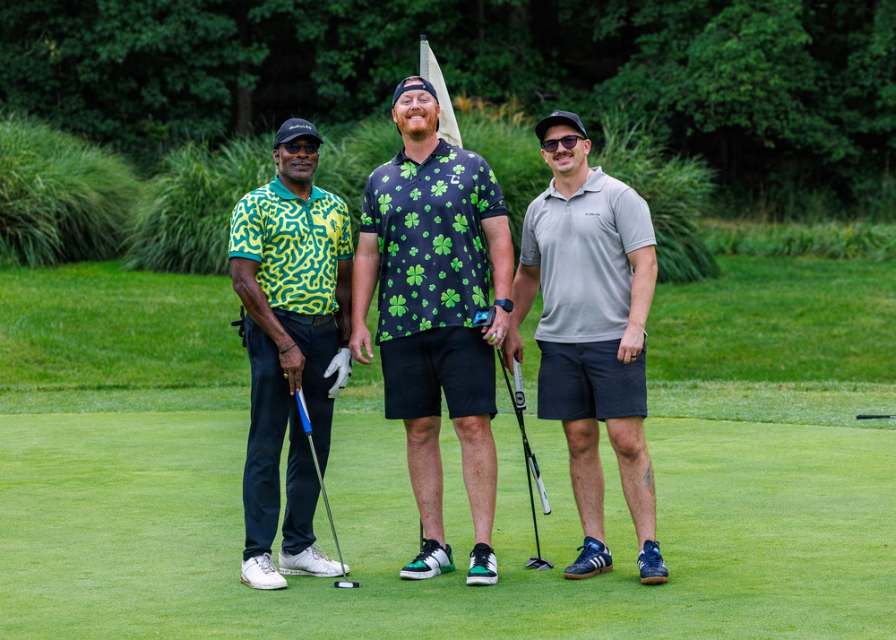 Three men are posing together on a golf course putting green, smiling at the camera. They are dressed in casual golf attire and two of them are holding putters. A golf flag is visible behind them, with lush green trees and ornamental grasses in the background.