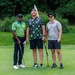 Three men are posing together on a golf course putting green, smiling at the camera. They are dressed in casual golf attire and two of them are holding putters. A golf flag is visible behind them, with lush green trees and ornamental grasses in the background.