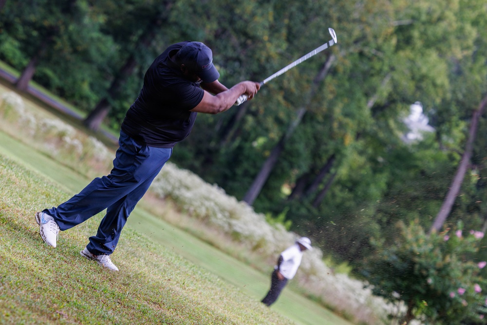 A golfer dressed in all black is captured mid-swing on a golf course, having just struck the ball with significant force. The follow-through position shows the club raised high behind the player's back, with visible turf debris from the impact. A second golfer in white clothing is visible in the blurred background, observing the shot.