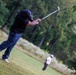 A golfer dressed in all black is captured mid-swing on a golf course, having just struck the ball with significant force. The follow-through position shows the club raised high behind the player's back, with visible turf debris from the impact. A second golfer in white clothing is visible in the blurred background, observing the shot.