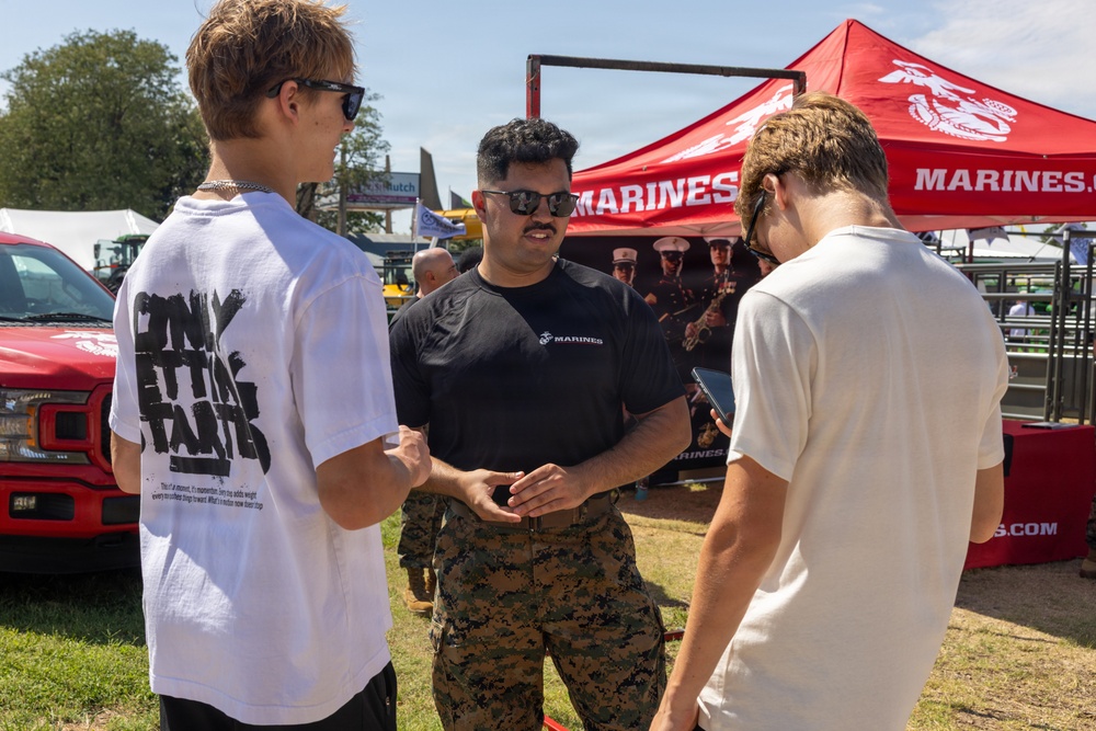A U.S. Marines recruiter in uniform is speaking with two young civilians at an outdoor recruiting booth or event. The recruiter, wearing a black Marines t-shirt and camouflage pants, appears to be engaged in conversation and possibly showing something on a device. The setting appears to be a fair or outdoor public event with a red Marines branded canopy tent visible in the background.