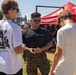 A U.S. Marines recruiter in uniform is speaking with two young civilians at an outdoor recruiting booth or event. The recruiter, wearing a black Marines t-shirt and camouflage pants, appears to be engaged in conversation and possibly showing something on a device. The setting appears to be a fair or outdoor public event with a red Marines branded canopy tent visible in the background.