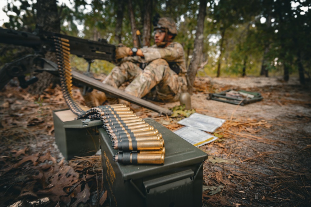 A soldier in camouflage uniform is positioned in a wooded area operating what appears to be a belt-fed machine gun. In the foreground, a linked ammunition belt is draped over a military-green ammunition can (ammo box). Documents or field manuals are visible on the ground nearby, suggesting a training or operational scenario.