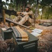 A soldier in camouflage uniform is positioned in a wooded area operating what appears to be a belt-fed machine gun. In the foreground, a linked ammunition belt is draped over a military-green ammunition can (ammo box). Documents or field manuals are visible on the ground nearby, suggesting a training or operational scenario.