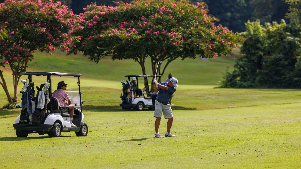 A golf course scene showing two players during a round of golf on a well-maintained fairway. One golfer is mid-swing executing a shot while another sits in a nearby golf cart observing. The background features blooming pink crape myrtle trees and lush green vegetation under bright sunlight.