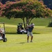 A golf course scene showing two players during a round of golf on a well-maintained fairway. One golfer is mid-swing executing a shot while another sits in a nearby golf cart observing. The background features blooming pink crape myrtle trees and lush green vegetation under bright sunlight.