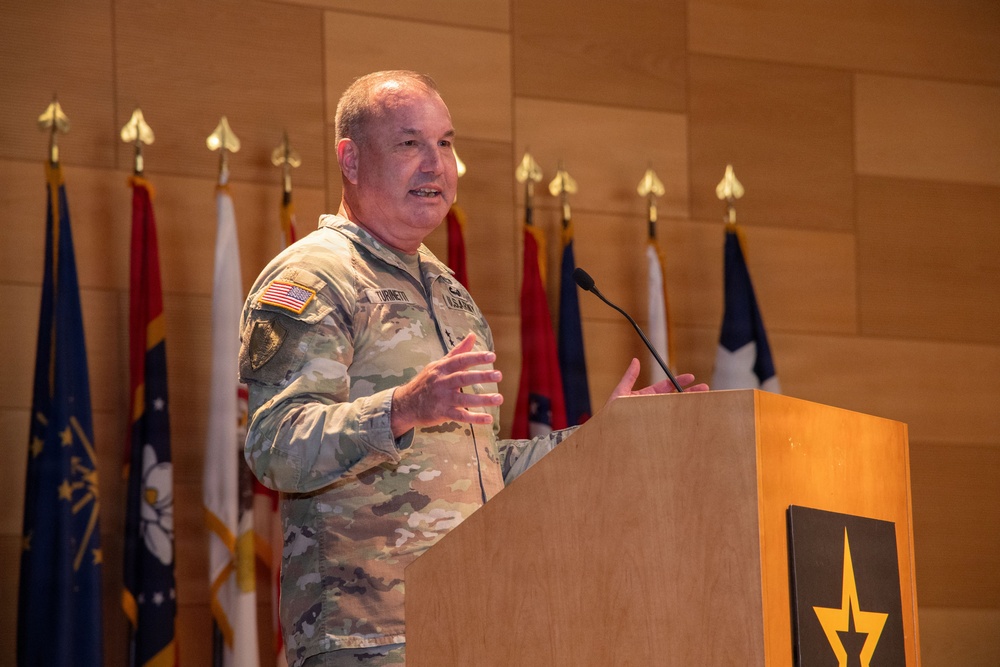 A U.S. Army officer in OCP (Operational Camouflage Pattern) uniform is delivering a speech at a wooden podium bearing the U.S. Army star logo. Multiple military and state flags are displayed on staffs in the background against a wood-paneled wall. The setting appears to be a formal military ceremony or official briefing.