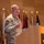 A U.S. Army officer in OCP (Operational Camouflage Pattern) uniform is delivering a speech at a wooden podium bearing the U.S. Army star logo. Multiple military and state flags are displayed on staffs in the background against a wood-paneled wall. The setting appears to be a formal military ceremony or official briefing.