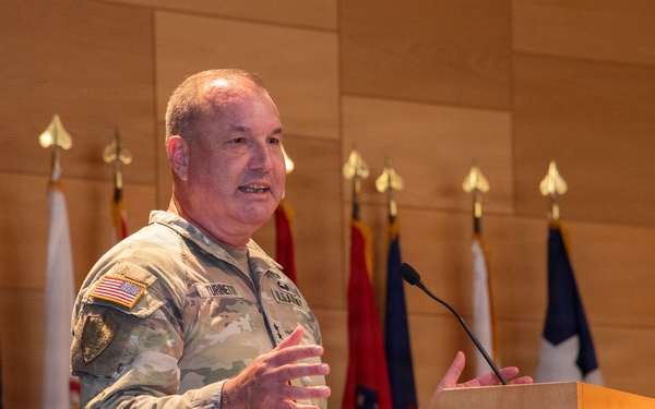 A U.S. Army officer in OCP (Operational Camouflage Pattern) uniform is delivering a speech at a wooden podium bearing the U.S. Army star logo. Multiple military and state flags are displayed on staffs in the background against a wood-paneled wall. The setting appears to be a formal military ceremony or official briefing.