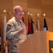 A U.S. Army officer in OCP (Operational Camouflage Pattern) uniform is delivering a speech at a wooden podium bearing the U.S. Army star logo. Multiple military and state flags are displayed on staffs in the background against a wood-paneled wall. The setting appears to be a formal military ceremony or official briefing.