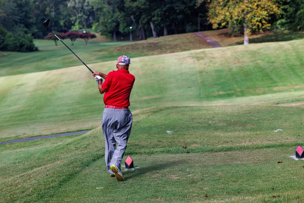 A golfer wearing a red polo shirt and gray pants has just completed a full follow-through swing on a golf course tee box. The player is photographed from behind, with the club raised high after impact. The well-maintained course features rolling green fairways and a tree-lined background.