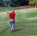 A golfer wearing a red polo shirt and gray pants has just completed a full follow-through swing on a golf course tee box. The player is photographed from behind, with the club raised high after impact. The well-maintained course features rolling green fairways and a tree-lined background.