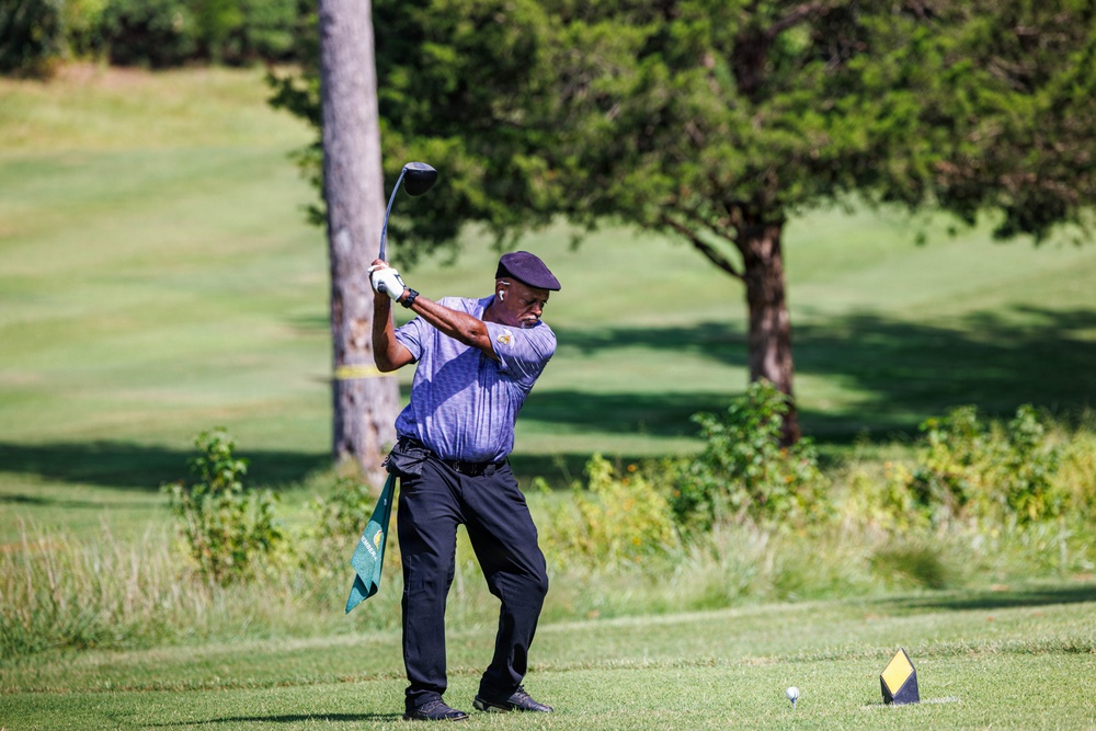 A golfer is captured mid-swing on a golf course, appearing to be at a tee box with a driver club raised in a follow-through position. The well-maintained course features lush green fairways, trees in the background, and a golf ball visible on a tee nearby. The image is sharp and well-lit with natural daylight.