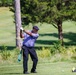 A golfer is captured mid-swing on a golf course, appearing to be at a tee box with a driver club raised in a follow-through position. The well-maintained course features lush green fairways, trees in the background, and a golf ball visible on a tee nearby. The image is sharp and well-lit with natural daylight.