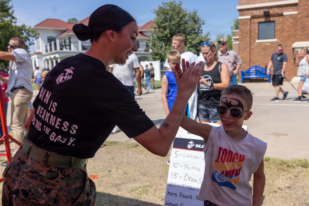 A female U.S. Marine in uniform gives a high-five to a young boy with Batman face paint at what appears to be a military community event or open house. The Marine is wearing a black t-shirt with the USMC Eagle, Globe, and Anchor emblem and the phrase 'Pain is Weakness Leaving the Body,' along with camouflage trousers. The outdoor event is taking place on a sunny day with families and other attendees visible in the background.