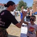A female U.S. Marine in uniform gives a high-five to a young boy with Batman face paint at what appears to be a military community event or open house. The Marine is wearing a black t-shirt with the USMC Eagle, Globe, and Anchor emblem and the phrase 'Pain is Weakness Leaving the Body,' along with camouflage trousers. The outdoor event is taking place on a sunny day with families and other attendees visible in the background.