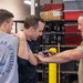 Three men are engaged in what appears to be a defensive tactics or self-defense training session in a gym or training facility. The central figure is examining or demonstrating a technique involving a blue training weapon (likely a rubber/foam training knife) held by the man on the right, while placing his hand on the shoulder of the man on the left. The setting includes cage-style fencing and storage racks typical of a martial arts or law enforcement training facility.