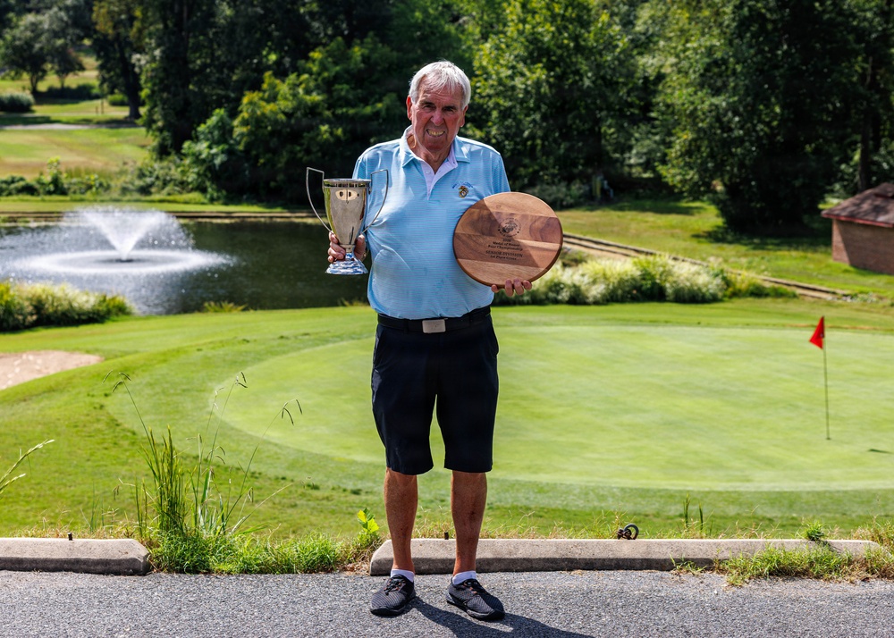 An elderly man stands on a golf course path holding a silver trophy cup in his left hand and a circular wooden plaque award in his right hand. He is smiling and dressed in golf attire, posing in front of a well-manicured green with a pond featuring a fountain and a red flag pin visible in the background. The setting and awards suggest he is celebrating a golf tournament victory or recognition.