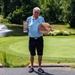 An elderly man stands on a golf course path holding a silver trophy cup in his left hand and a circular wooden plaque award in his right hand. He is smiling and dressed in golf attire, posing in front of a well-manicured green with a pond featuring a fountain and a red flag pin visible in the background. The setting and awards suggest he is celebrating a golf tournament victory or recognition.