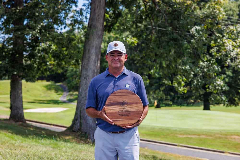 A middle-aged man stands on a golf course holding a large circular wooden award plaque. He is dressed in golf attire including a blue polo shirt, light gray pants, and a white cap. The setting is a well-maintained golf course with trees, a sand bunker, and a green visible in the background.