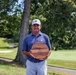 A middle-aged man stands on a golf course holding a large circular wooden award plaque. He is dressed in golf attire including a blue polo shirt, light gray pants, and a white cap. The setting is a well-maintained golf course with trees, a sand bunker, and a green visible in the background.