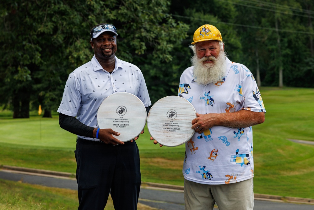 Two men are posing outdoors on a golf course, each holding a circular wooden award plaque. The plaques are from the 2026 Medal of Honor Base Championship — one for Men's Division 1st Place Net and the other for Senior Division 1st Place Net. Both men are smiling, dressed in golf attire, with a lush green course visible in the background.