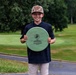 A young boy smiles proudly while holding a large circular wooden award plaque on what appears to be a golf course. The plaque reads '2026 Medal of Honor Base Championship, Junior Division, 1st Place.' He is dressed in a navy polo shirt and camouflage cap, standing on a cart path with a green fairway and trees visible in the background.