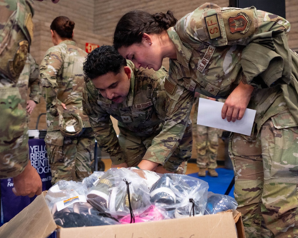 Two U.S. Air Force personnel in OCP (Operational Camouflage Pattern) uniforms are examining the contents of a cardboard box filled with plastic-wrapped items, likely shoes or athletic gear. One service member is holding a document or paper while both lean over the box, suggesting an inventory or distribution activity. The setting appears to be an indoor gymnasium or community center, with a 'Yellow Ribbon Reintegration Program' banner visible in the background.