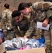 Two U.S. Air Force personnel in OCP (Operational Camouflage Pattern) uniforms are examining the contents of a cardboard box filled with plastic-wrapped items, likely shoes or athletic gear. One service member is holding a document or paper while both lean over the box, suggesting an inventory or distribution activity. The setting appears to be an indoor gymnasium or community center, with a 'Yellow Ribbon Reintegration Program' banner visible in the background.