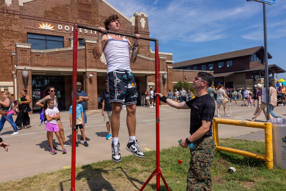 A young man in a white tank top and floral shorts performs pull-ups on a red portable pull-up bar at what appears to be an outdoor public event or festival. A person in Marine Corps camouflage uniform stands nearby, likely counting or supervising the exercise. The event is taking place in a parking lot area with a brick building labeled 'Domestic' visible in the background, surrounded by a crowd of onlookers.