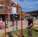 A young man in a white tank top and floral shorts performs pull-ups on a red portable pull-up bar at what appears to be an outdoor public event or festival. A person in Marine Corps camouflage uniform stands nearby, likely counting or supervising the exercise. The event is taking place in a parking lot area with a brick building labeled 'Domestic' visible in the background, surrounded by a crowd of onlookers.
