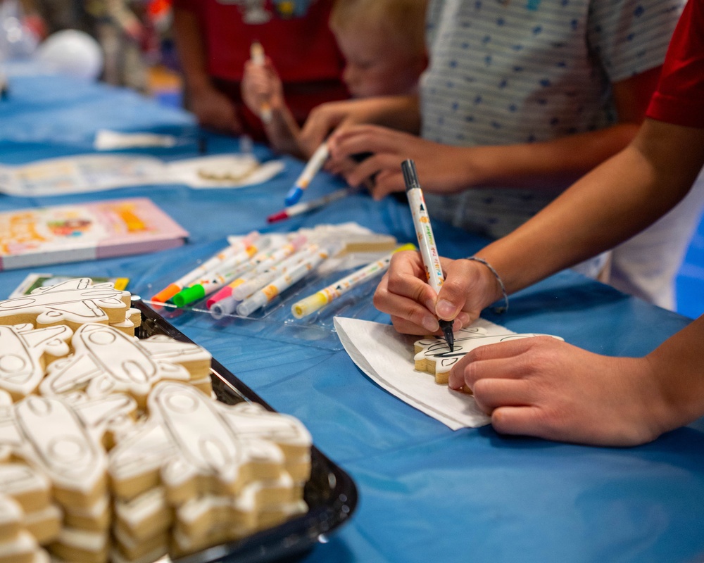 Children are participating in a cookie decorating activity at a table covered with a blue tablecloth. They are using food-safe markers to draw designs on white-frosted sugar cookies. A tray of undecorated cookies is visible in the foreground, along with multiple packets of colorful markers.