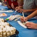 Children are participating in a cookie decorating activity at a table covered with a blue tablecloth. They are using food-safe markers to draw designs on white-frosted sugar cookies. A tray of undecorated cookies is visible in the foreground, along with multiple packets of colorful markers.