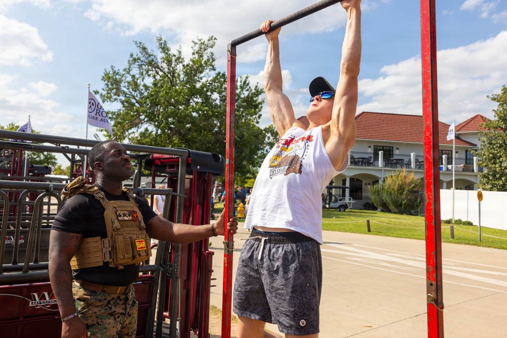 A muscular man in a white sleeveless shirt, black cap, and blue sunglasses performs pull-ups on a red metal pull-up bar station outdoors. A man wearing a black t-shirt and tan tactical/plate carrier vest with a 'SAR' patch observes and steadies the equipment. The setting appears to be an outdoor public event or fair, with a building, trees, and a flag visible in the background.